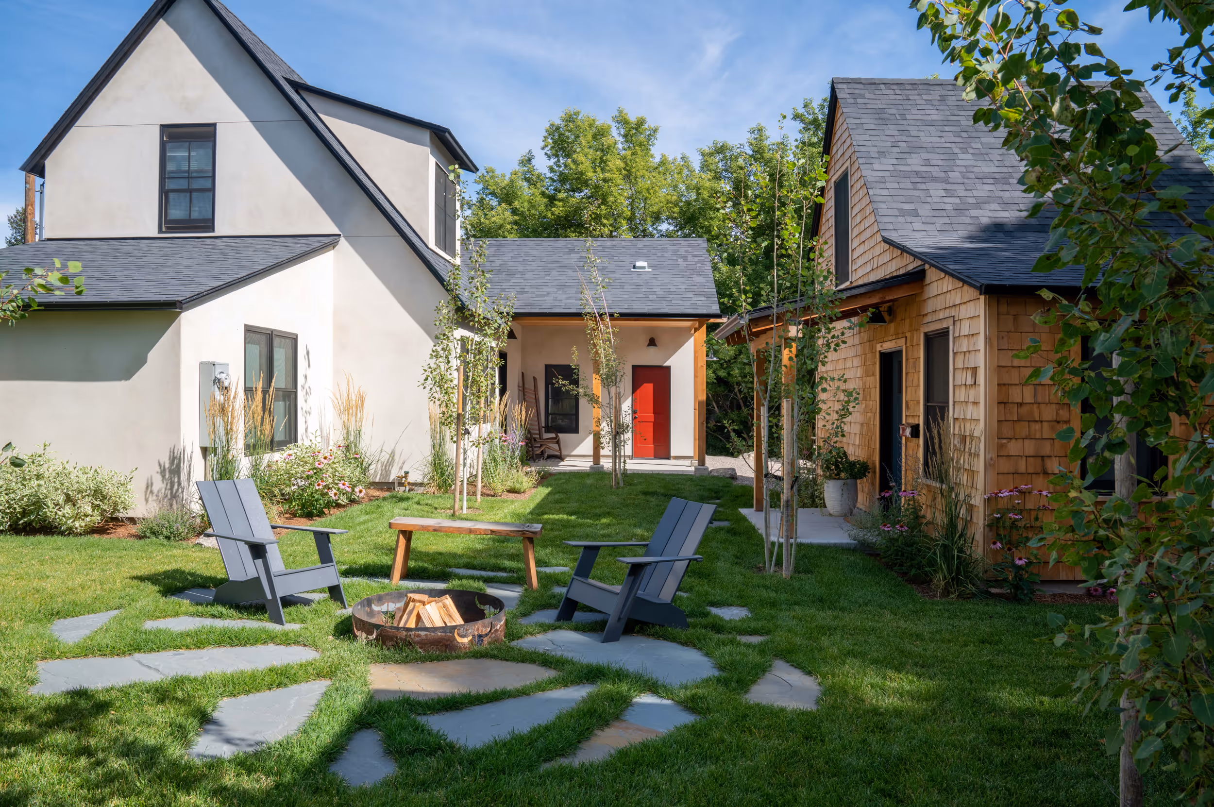 A yard with an outdoor fire place in the foreground, and a wooden garage, and charming, minimalistic house with a red door in the background
