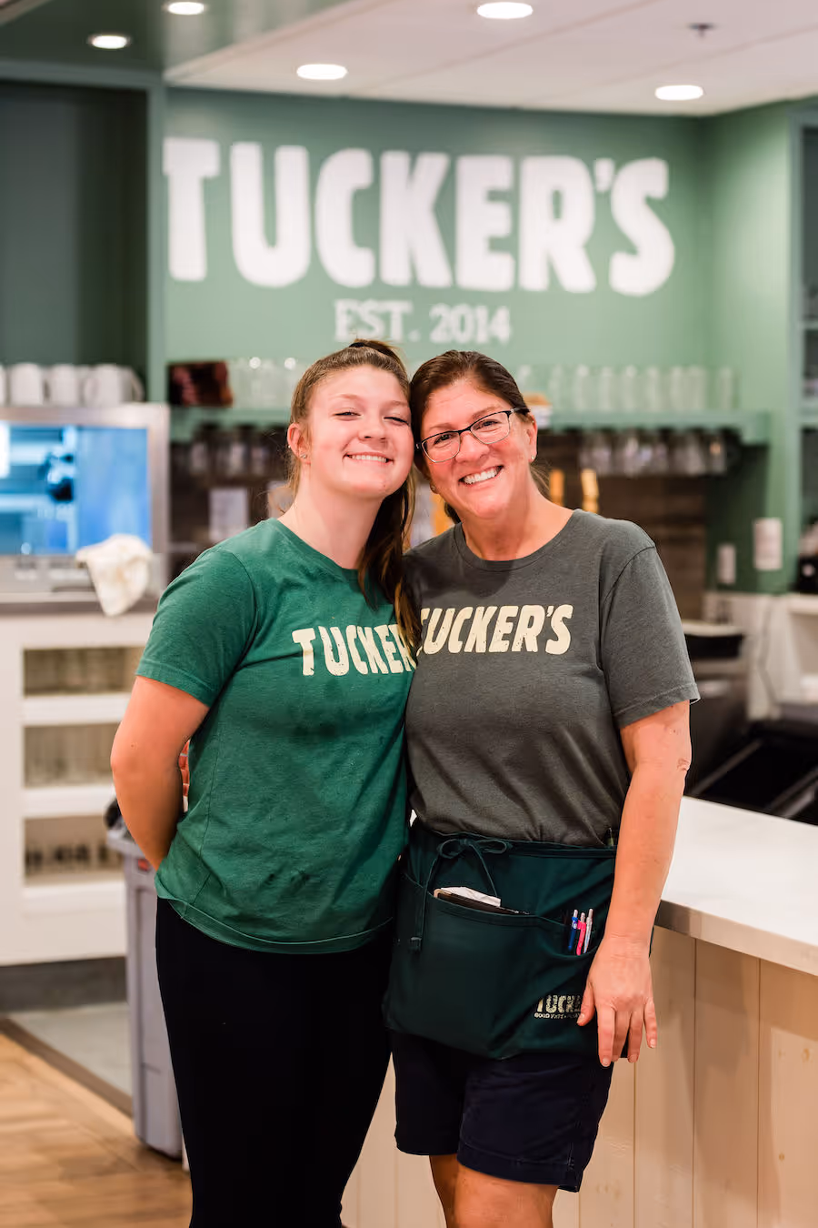 Two people wearing matching "Tucker's" shirts, standing and smiling inside a restaurant with "Tucker's" sign in the background.