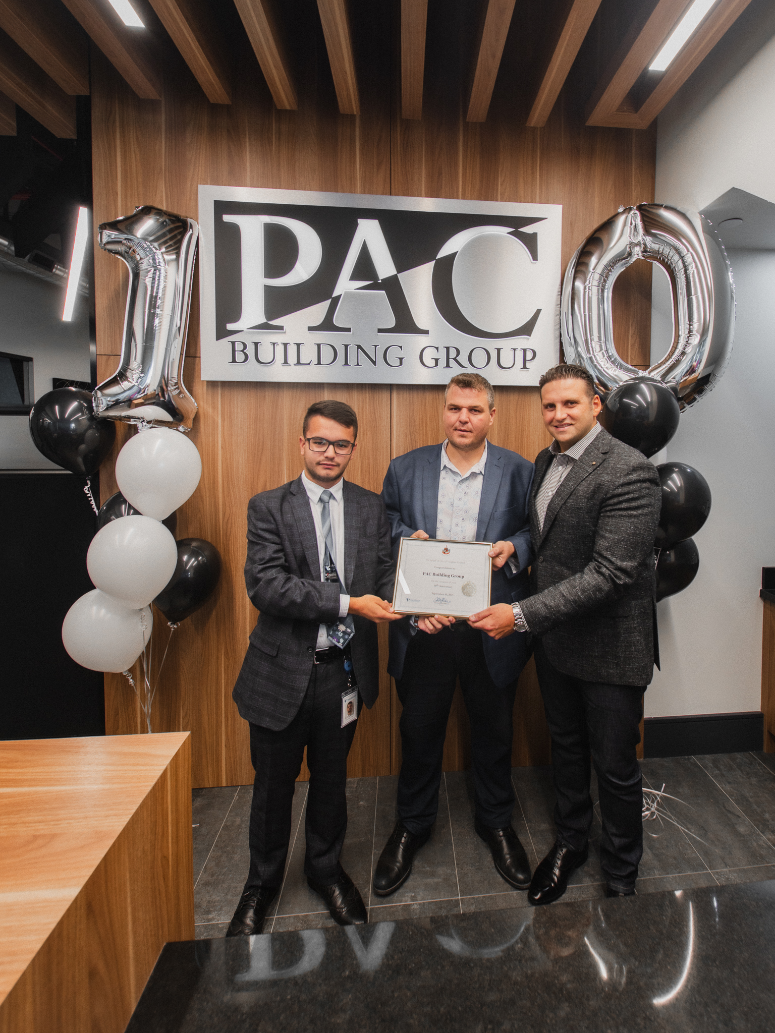 La direction de PAC Building Group tient un certificat du 10e anniversaire devant le logo de PAC