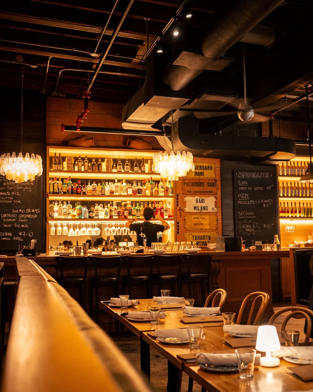 Warmly lit bar with an illuminated bottle display and Bari, Milano, Toronto signage