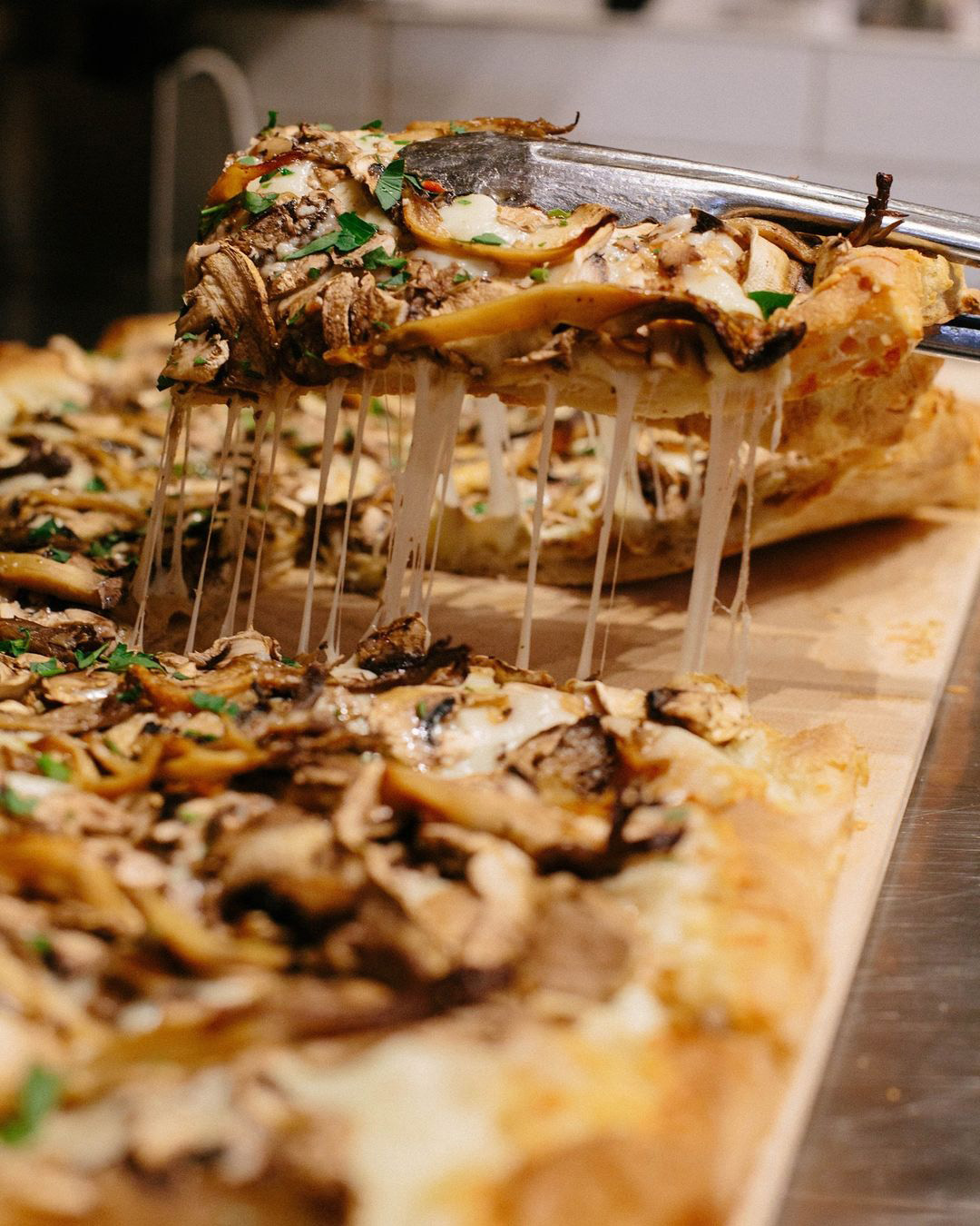 Pizza slice lifted from the wooden display board, melted cheese stretching down, topped with mushrooms