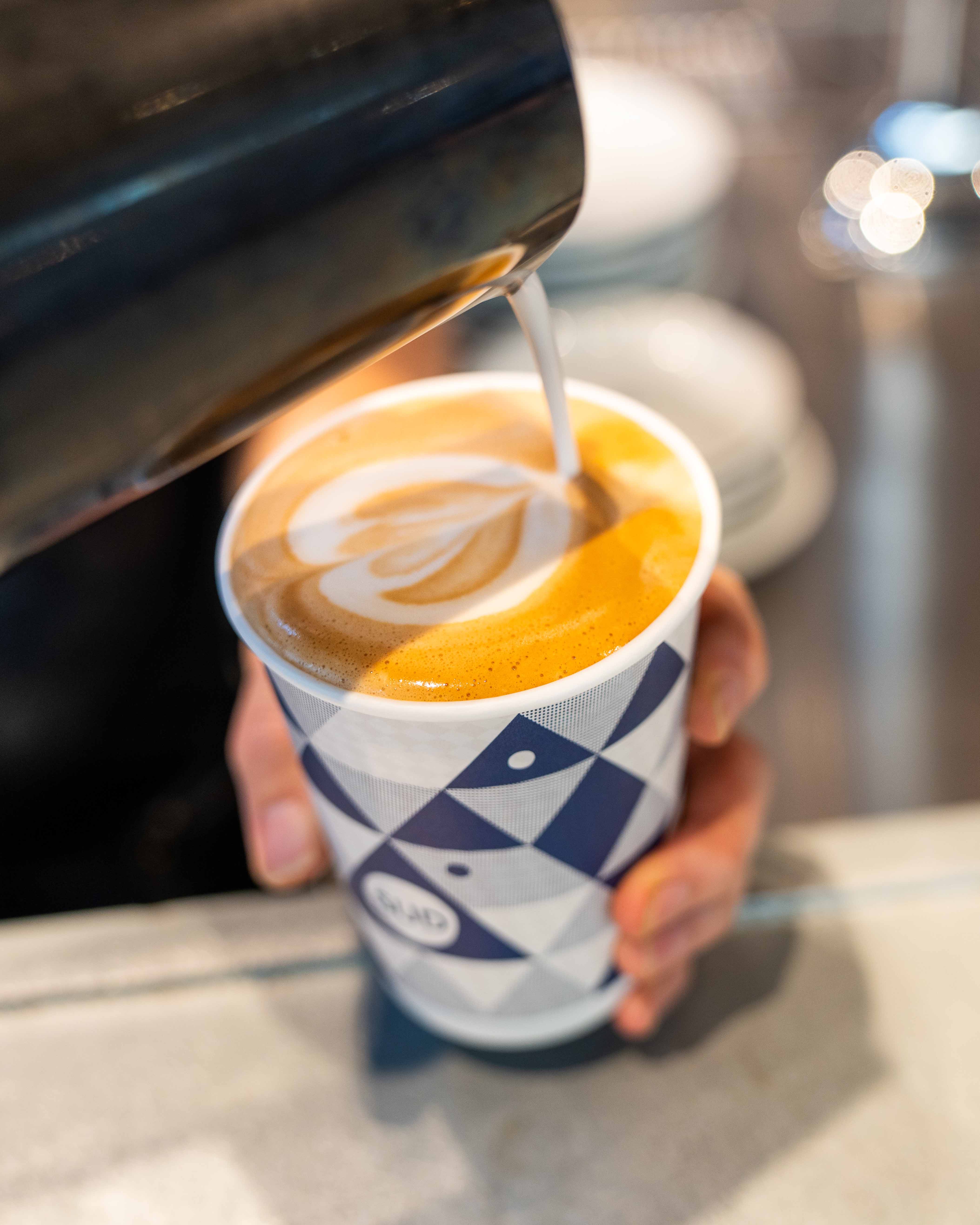 Barista pouring latte art into a paper cup stamped with the Sud logo