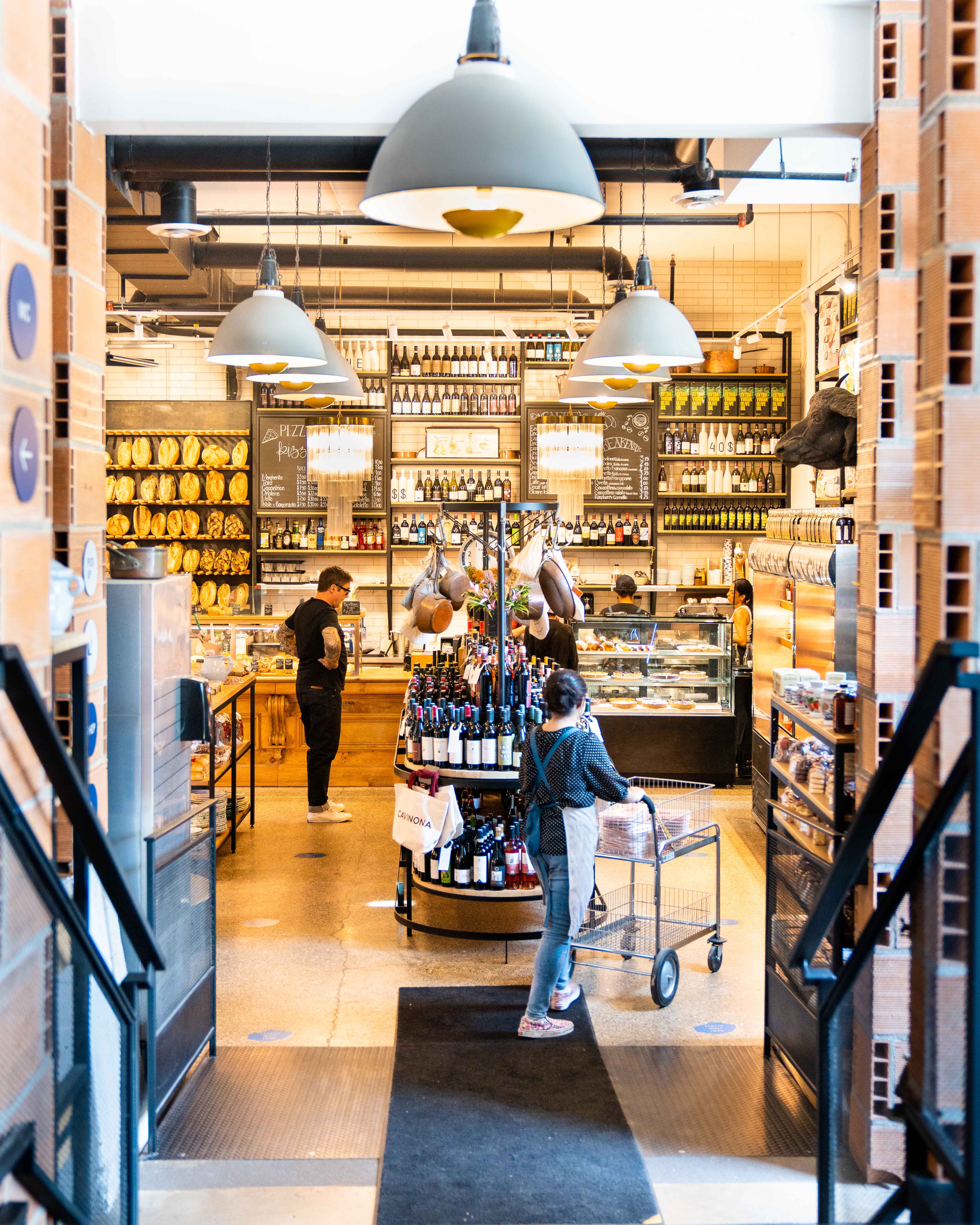 View into the grocer with wine shelves, bread racks, and a shopper pushing a cart