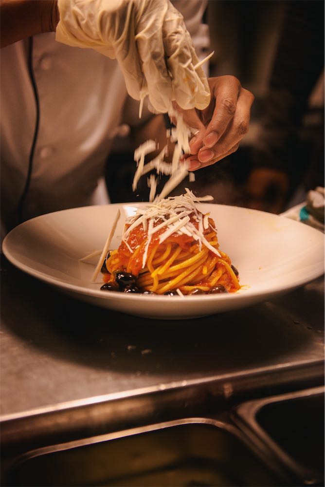 Chef sprinkling ricotta salata over spaghetti with tomato sauce and olives