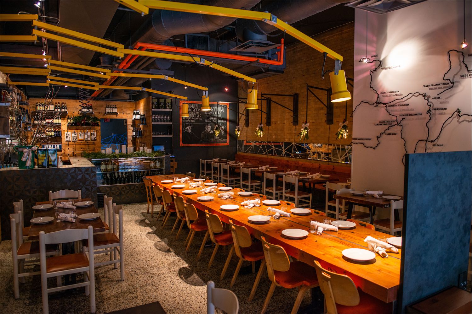 Private dining room with a long wood table set for a group, wine map mural and yellow ceiling pipes overhead