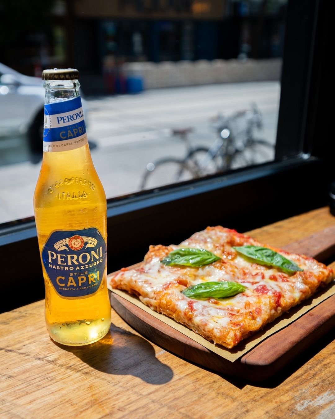 Peroni Capri beer bottle beside a slice of Margherita pizza by the window