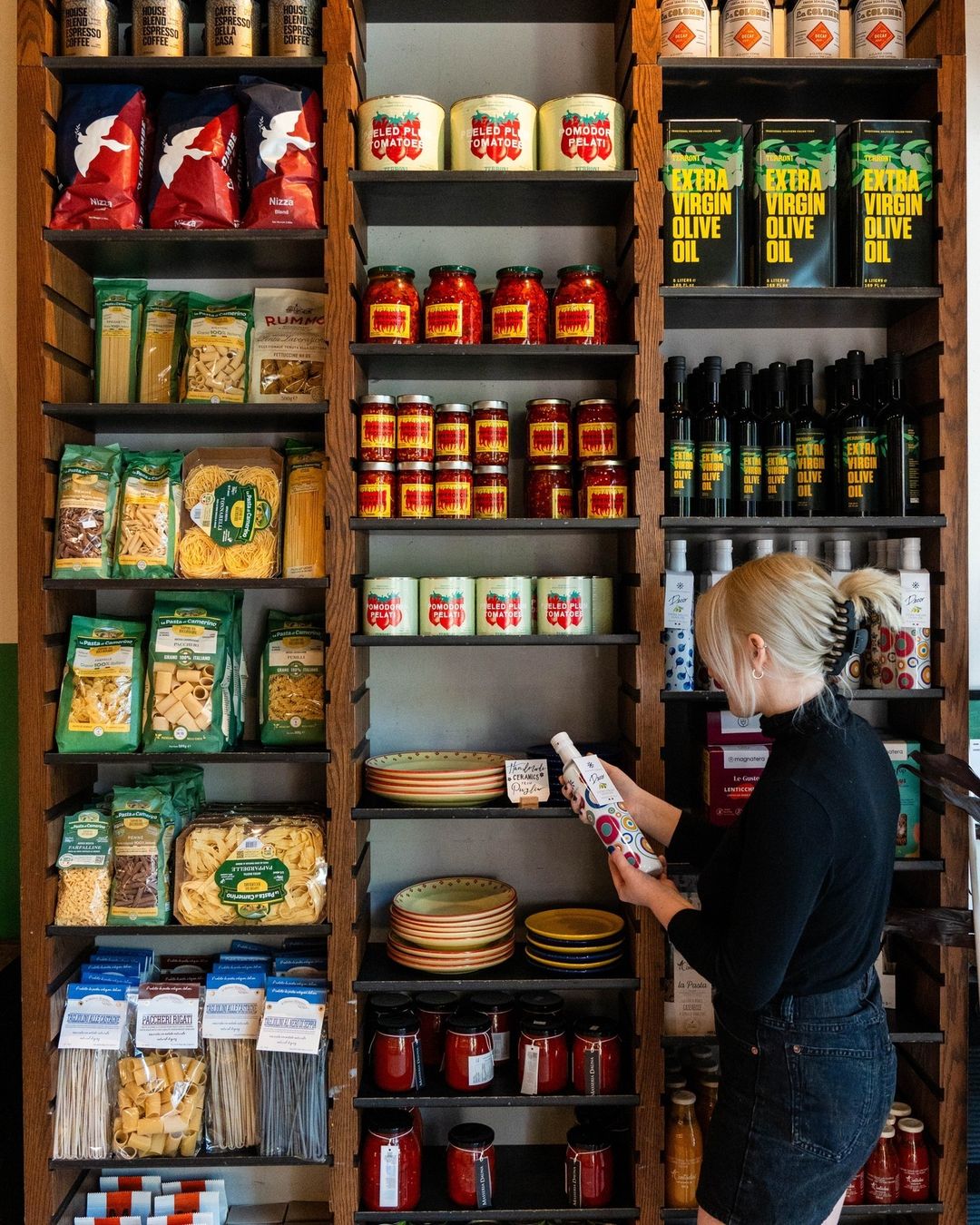 Woman shopping grocery shelves stocked with pasta, tomatoes, and olive oil tins