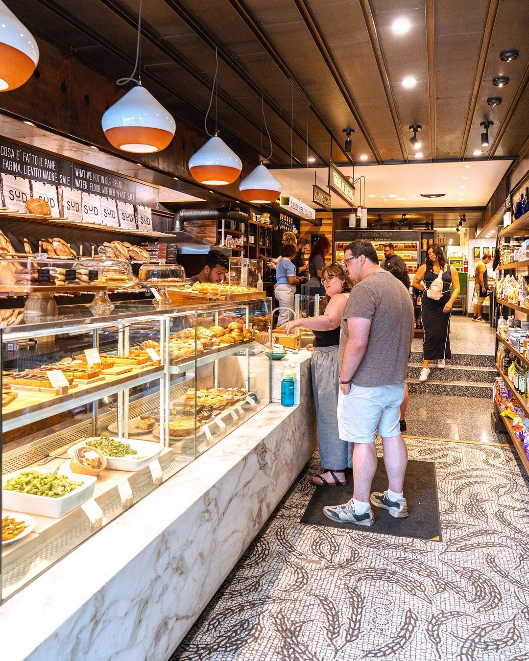 Customers at the marble pastry counter beneath bread shelves and pendant lights