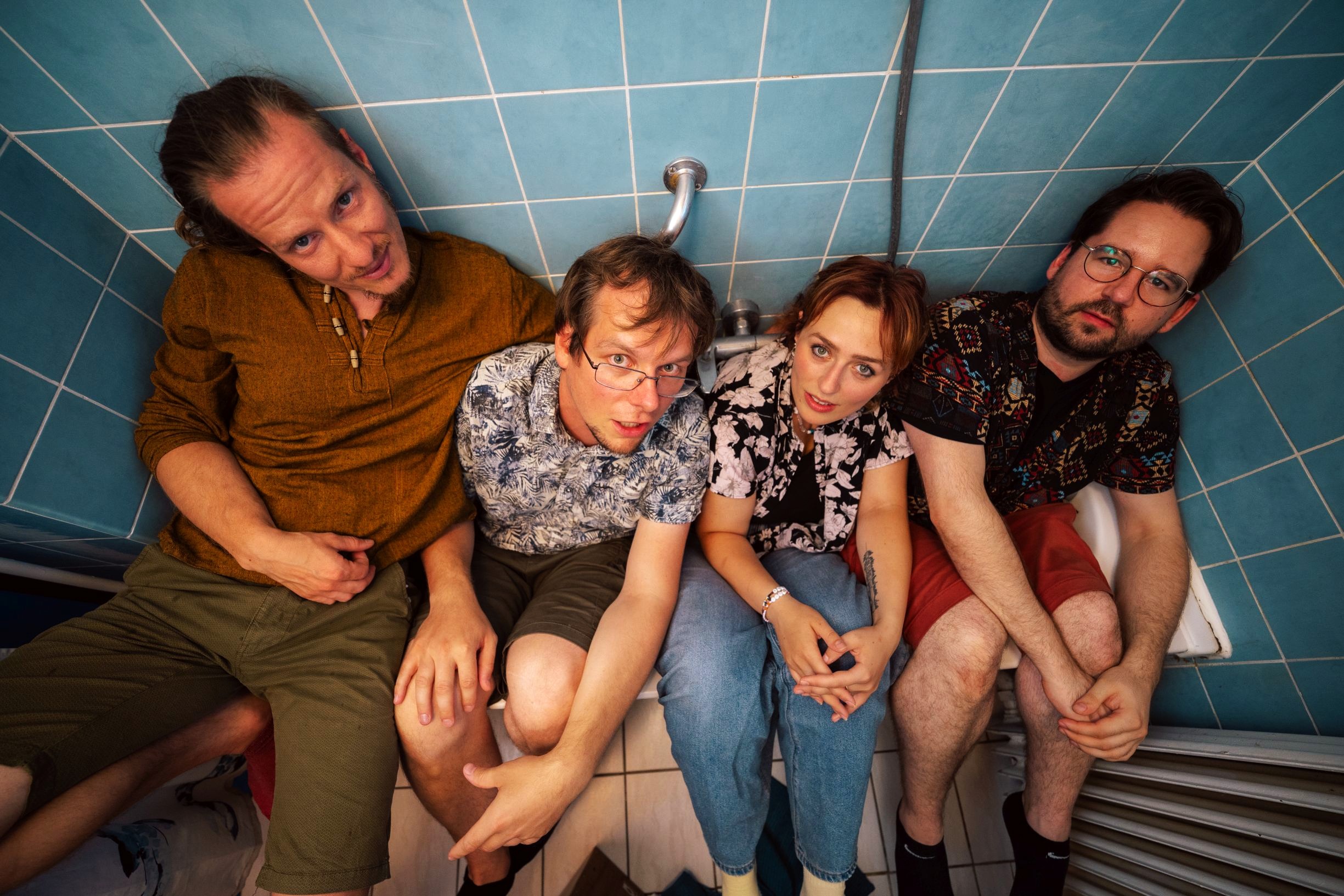 Four people outdoors with two men supporting a woman sitting on a white chair while another man lies under the chair on grass with green foliage in the background.