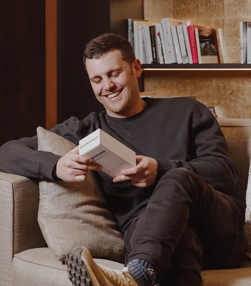 Happy man relaxing on a sofa, smiling while reading information from his Dispensed delivery box at home.