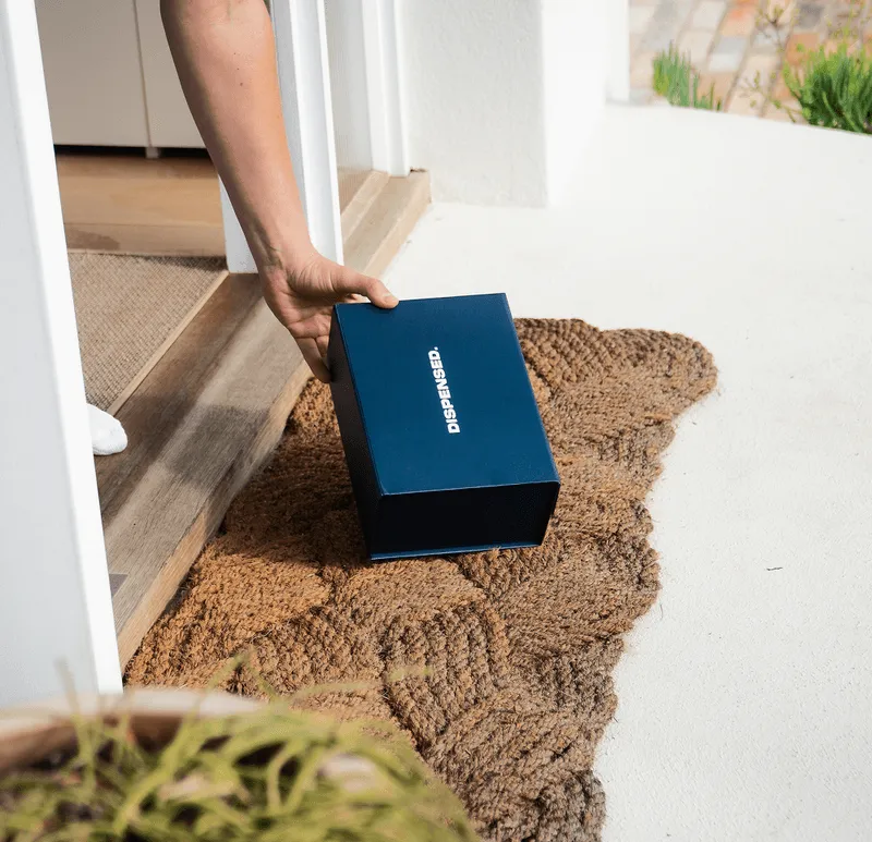 Close-up of a Dispensed delivery box being placed on a front doorstep, sitting on a door mat and being picked up by a patient.