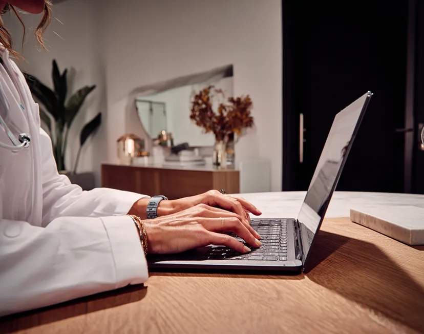 A clinician in a lab coat is sitting at their desk typing on their computer in a well lit home office environment.