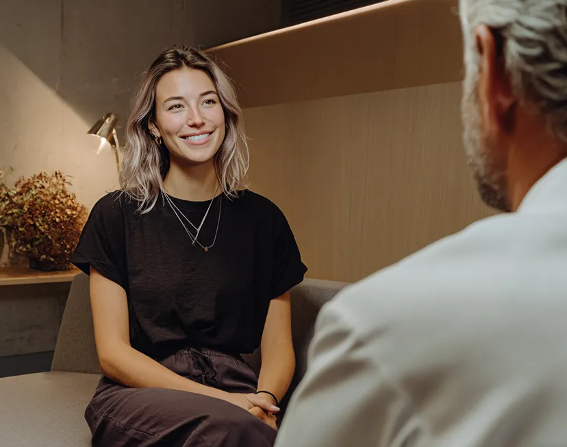 A smiling woman is sitting across from a clinician in a lab coat in a well lit office environment.