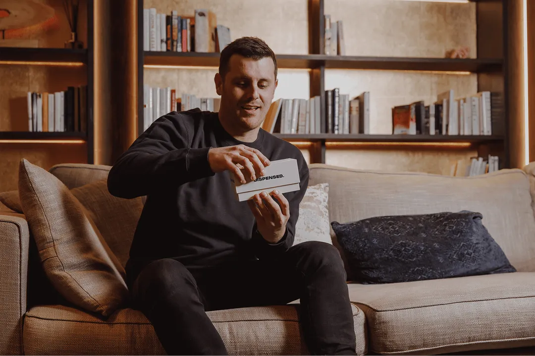 Man sitting comfortably on a couch, smiling as he opens a Dispensed medical delivery box in a warmly lit living room.