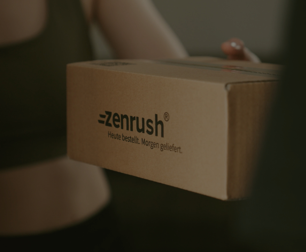 Hand hält Versandkarton mit Zenrush® Logo und Slogan „Heute bestellt. Morgen geliefert.“
