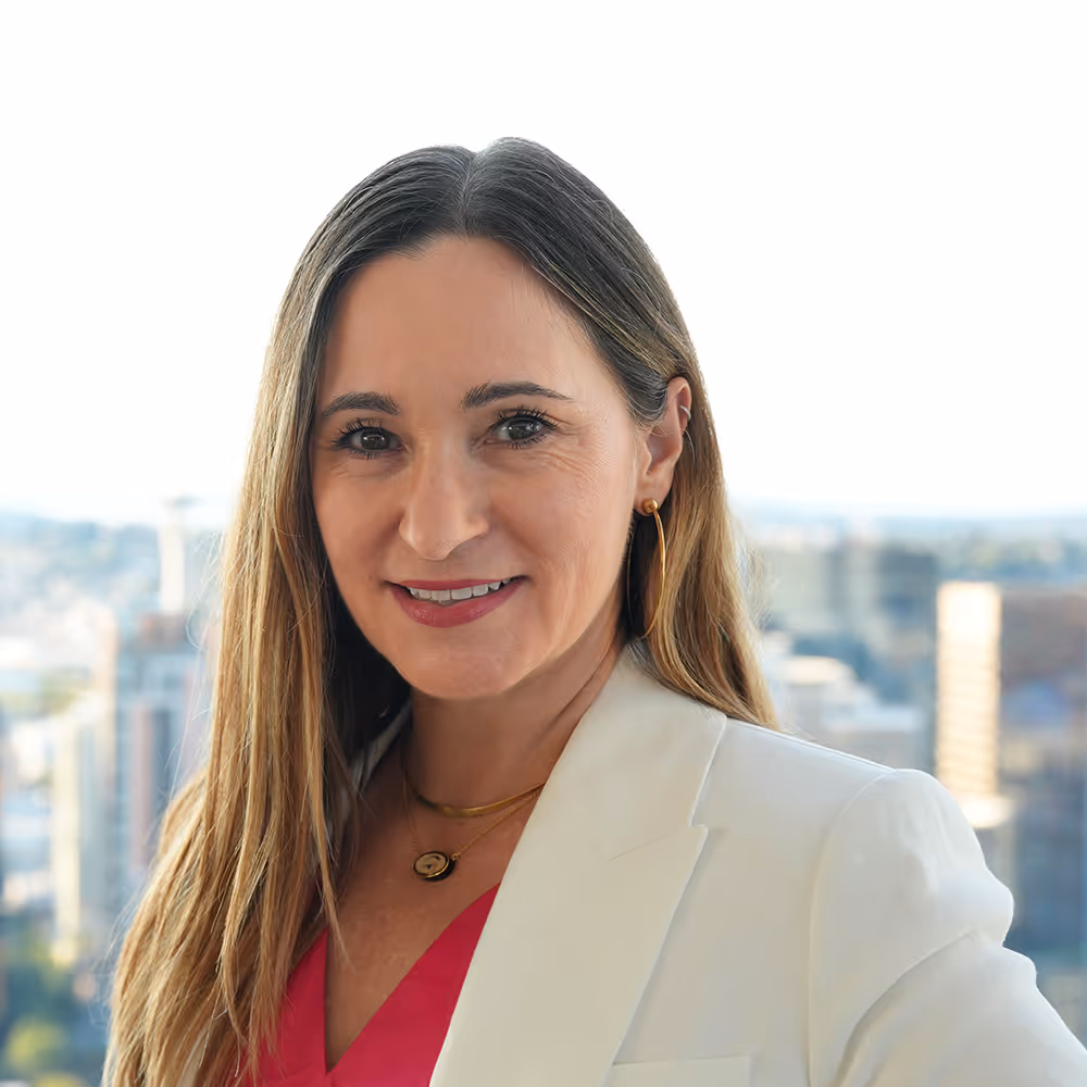 A photo of Kalie, with long brown hair wearing a white blazer and pink top, smiling against a cityscape background.