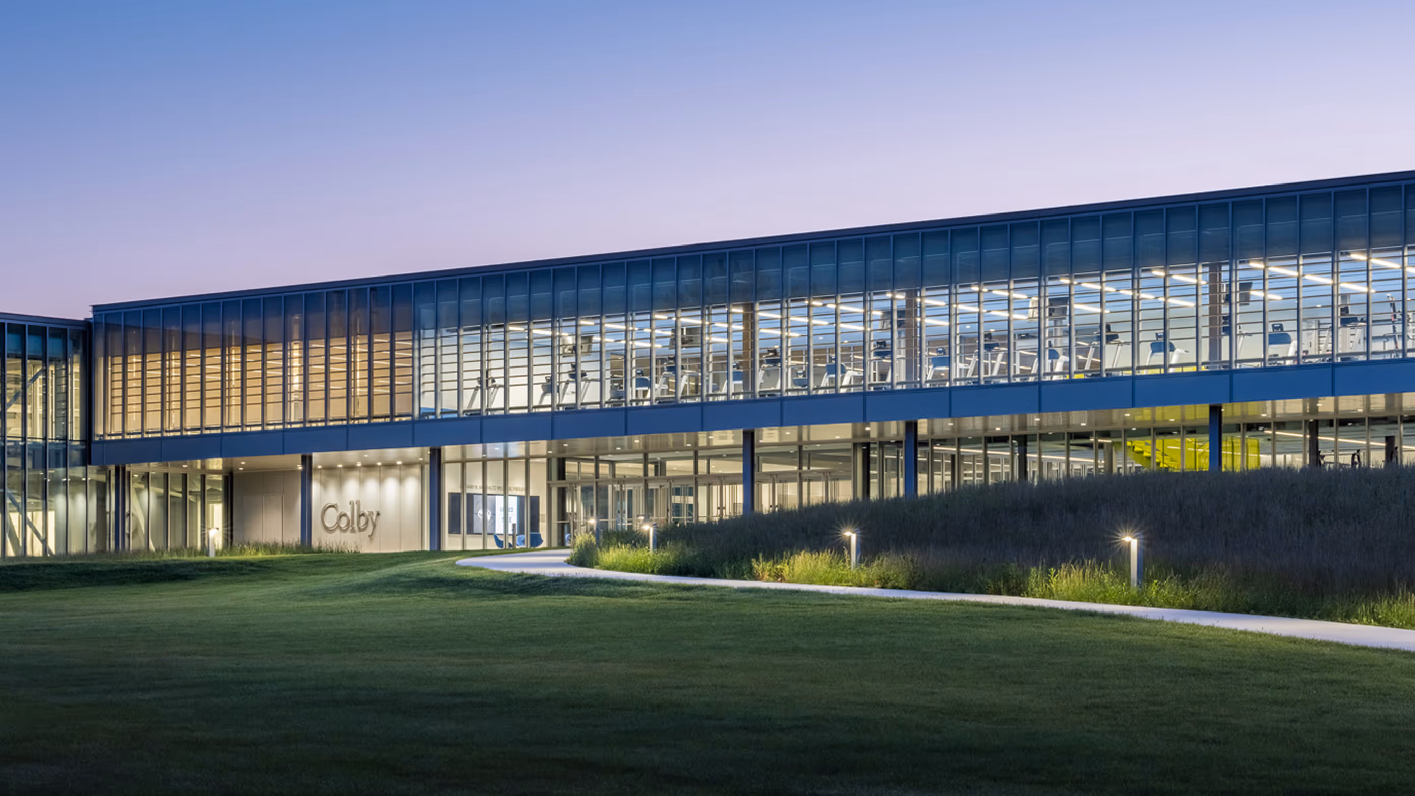 Modern glass-fronted building at dusk with illuminated interior and a pathway curving through a green lawn in the foreground.