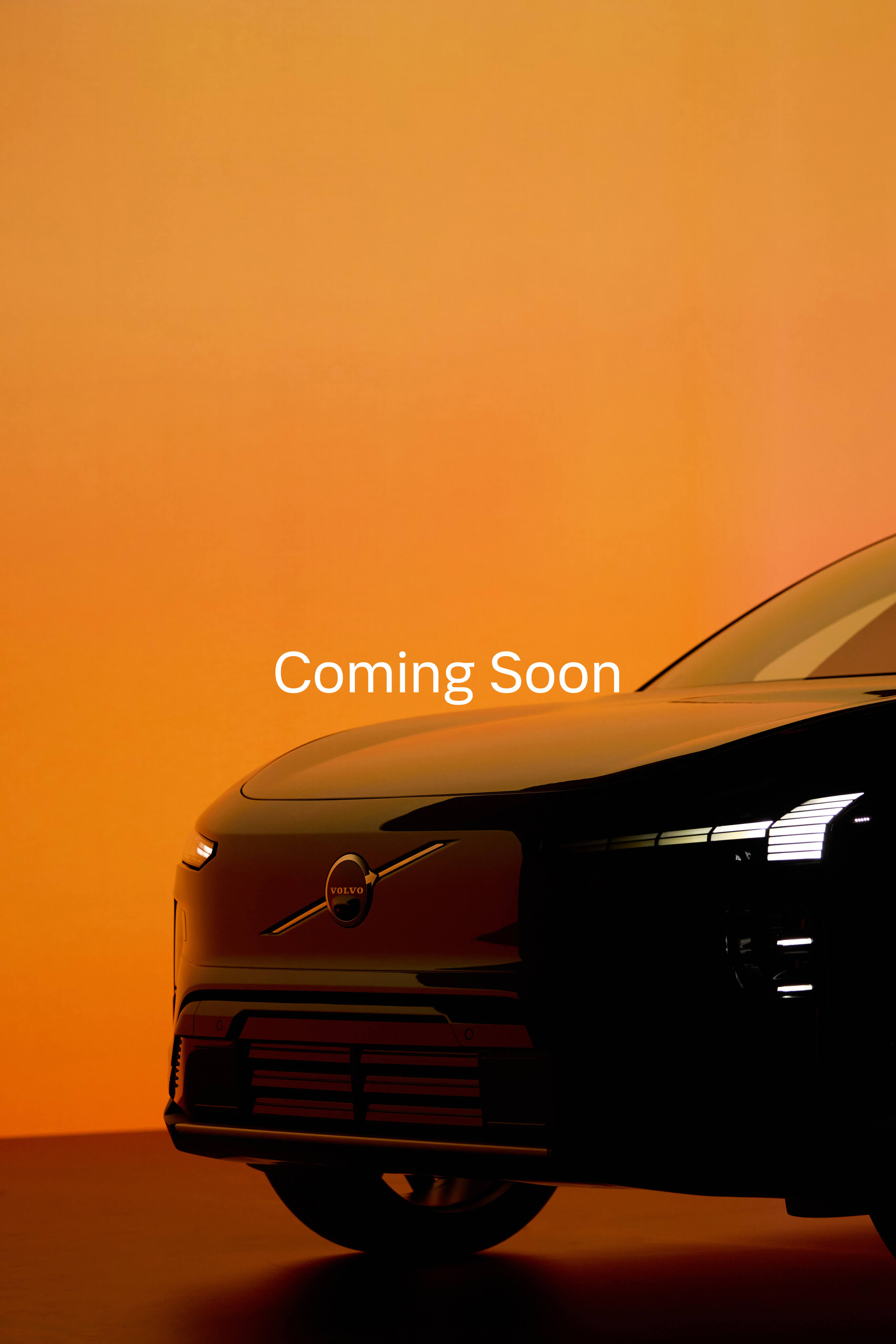 Front side view of a black Volvo car with headlights on against an orange background and the text 'Coming Soon.'
