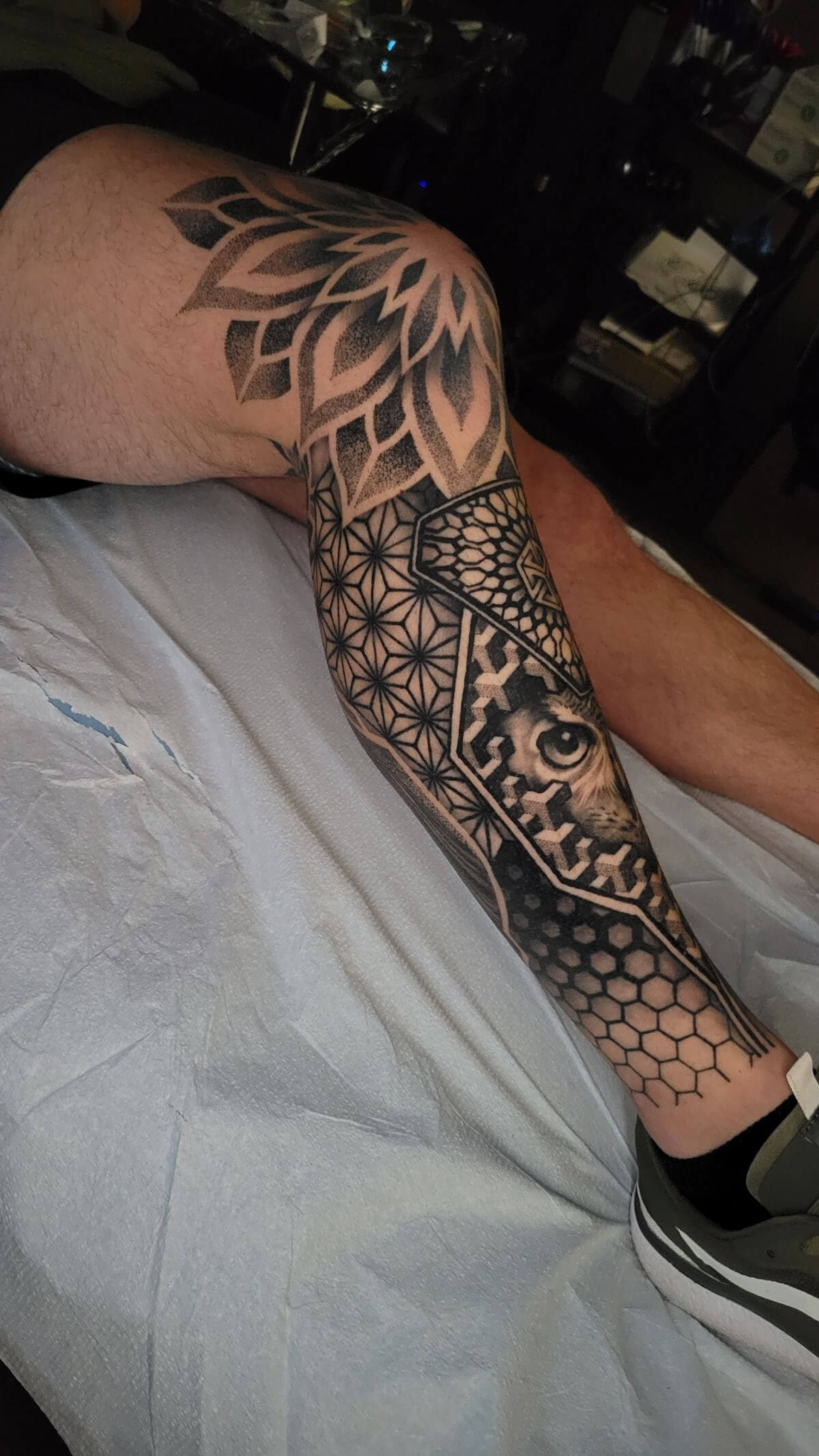 Black and grey geometric sleeve tattoo on leg featuring a stylized eye, honeycomb, and flower petal patterns by Jason Mims.