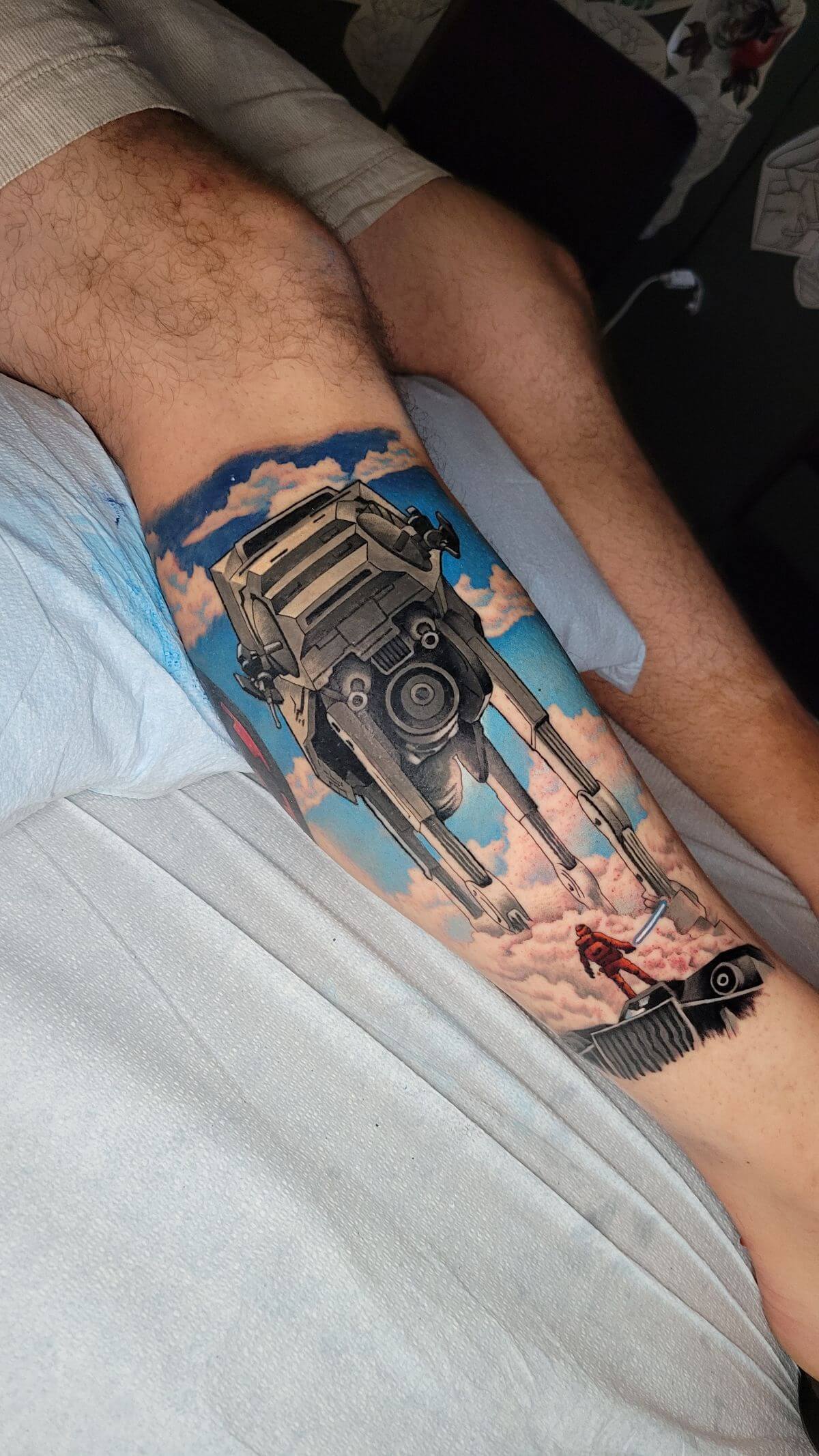 Tattoo on a leg depicting a Star Wars AT-AT walker descending through clouds toward a person in red armor holding a blue lightsaber.