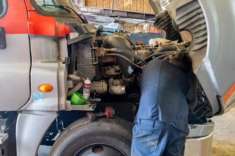 Fleet maintenance technician working under the hood of a semi-truck in a service bay, performing diagnostics and mechanical repairs to keep the vehicle road-ready.