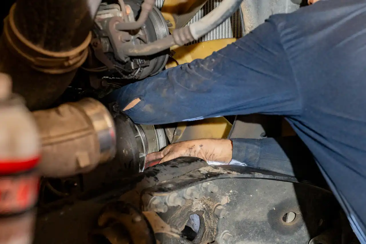 Fleet maintenance technician working under the hood of a semi-truck in a service bay, performing diagnostics and mechanical repairs to keep the vehicle road-ready.