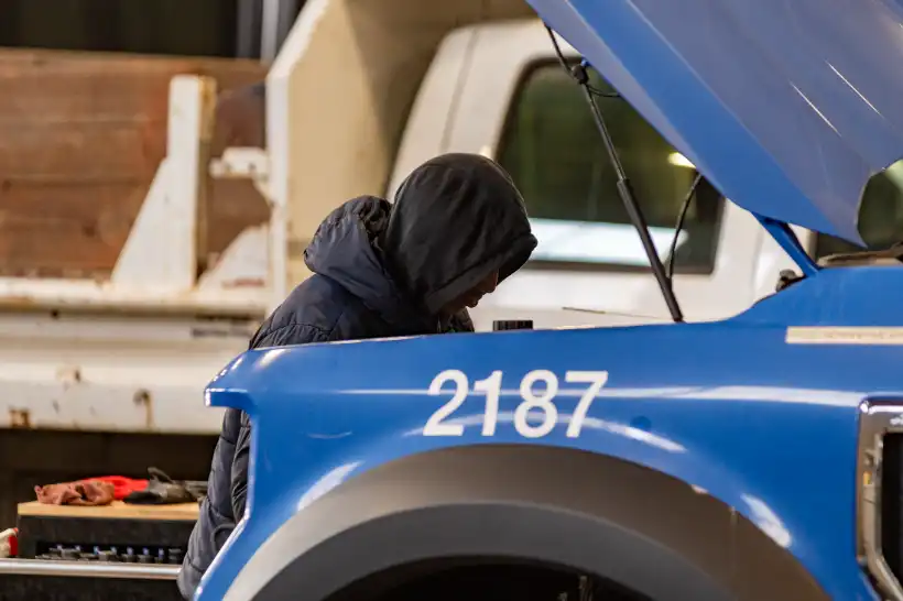 Fleet maintenance technician in hoodie inspects engine bay of blue service truck labeled 2187.