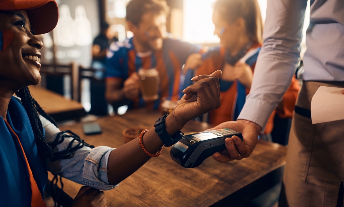 Image of people at a restaurant. There are two people blurred in the back talking to each other while sitting at a table. The focus is on a woman sitting at the same table who is using her smart watch to pay at a mobile checkout system a man is holding.