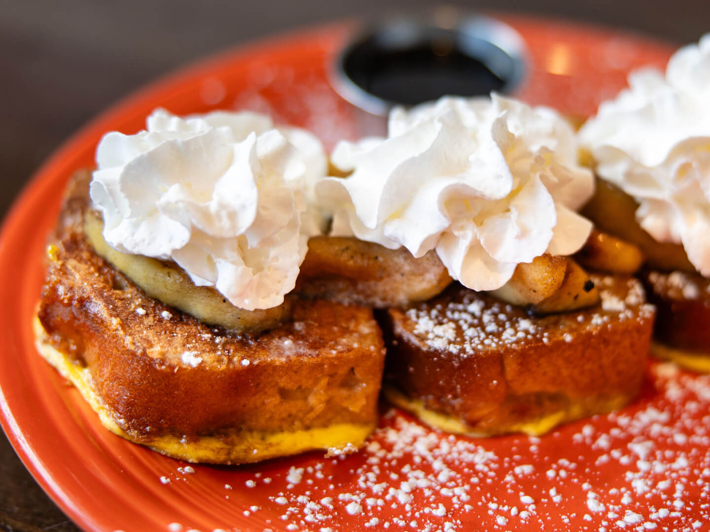 Three pieces of French toast topped with banana slices, whipped cream, and powdered sugar on an orange plate.