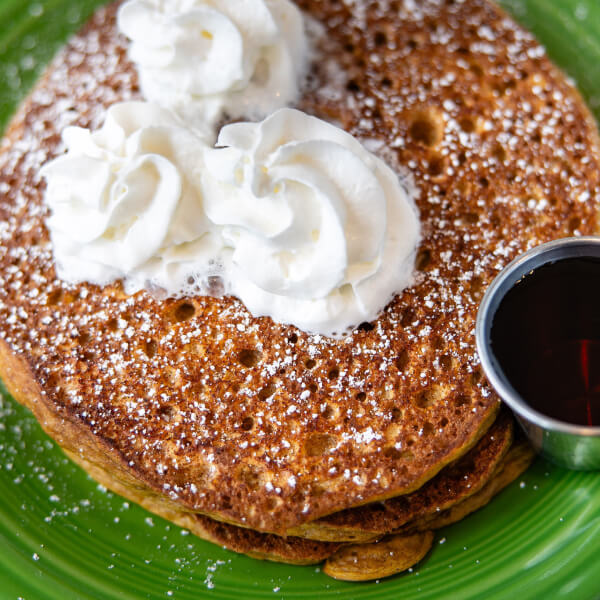 Two pancakes topped with whipped cream and powdered sugar, served with a side of syrup on a green plate.