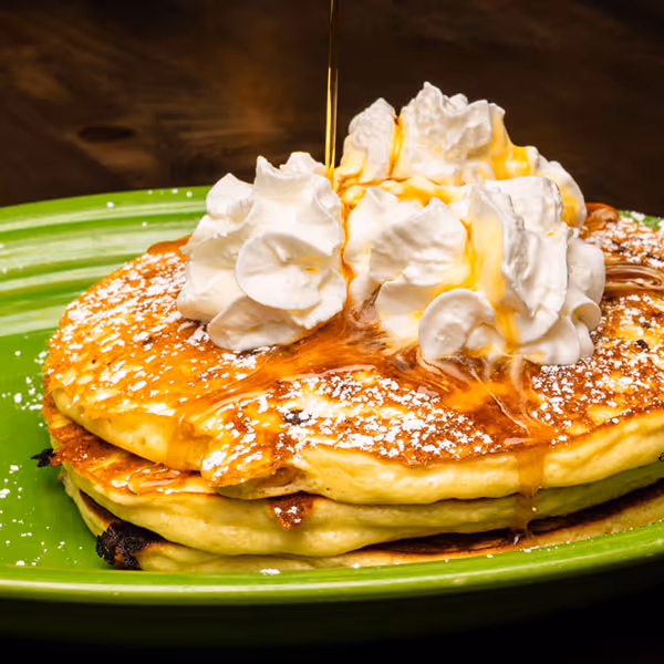 Stack of pancakes topped with whipped cream and powdered sugar, being drizzled with syrup on a green plate.