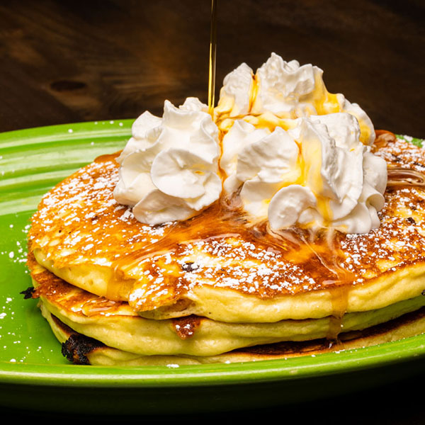 Stack of pancakes topped with whipped cream and powdered sugar, being drizzled with syrup on a green plate.