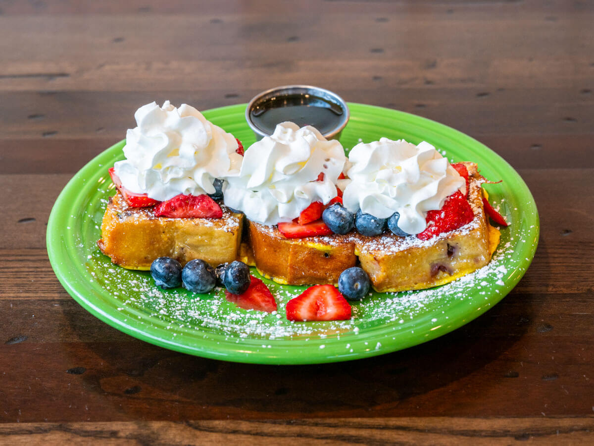 Three slices of French toast topped with whipped cream, strawberries, blueberries, and powdered sugar on a green plate.