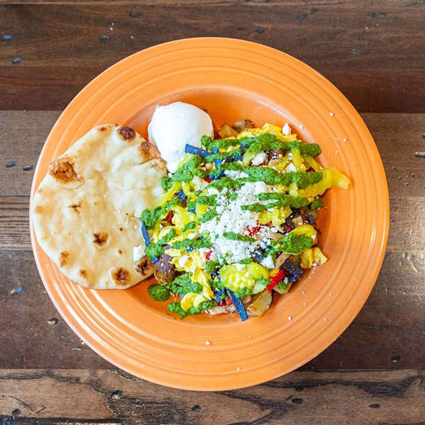 An orange plate with flatbread, a scoop of white spread, and a vegetable scramble topped with sauces and cheese.