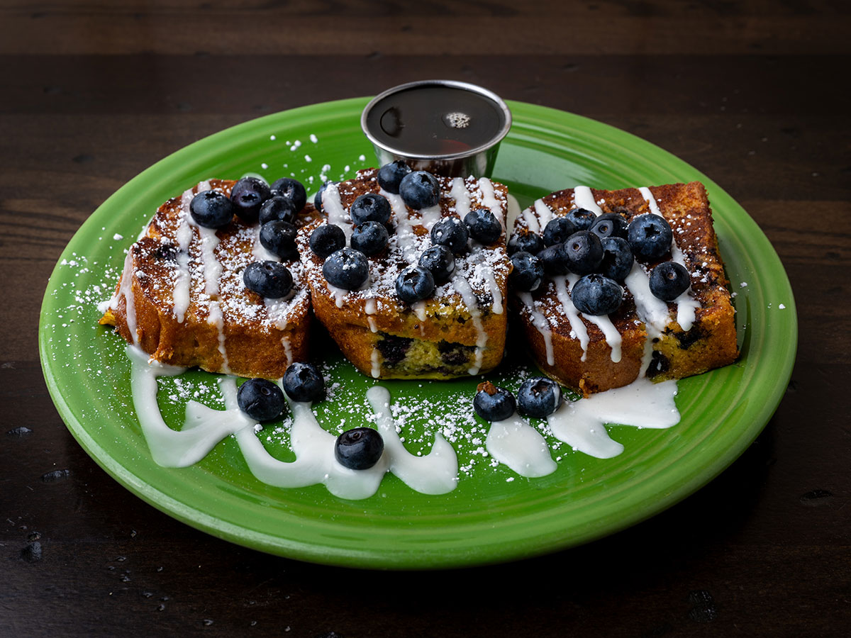 Three slices of French toast topped with blueberries, powdered sugar, and icing on a green plate with syrup on the side.