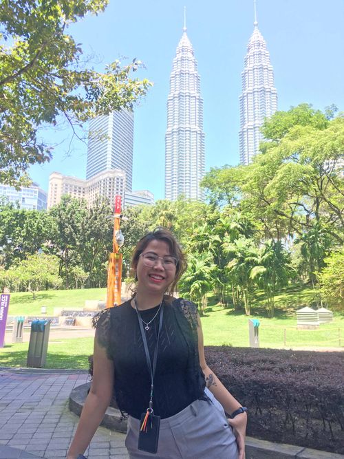 A picture of Depe wearing a black top at the KLCC Garden, Kuala Lumpur, the Petronas Twin Towers behind her with a blue sky as a backdrop.