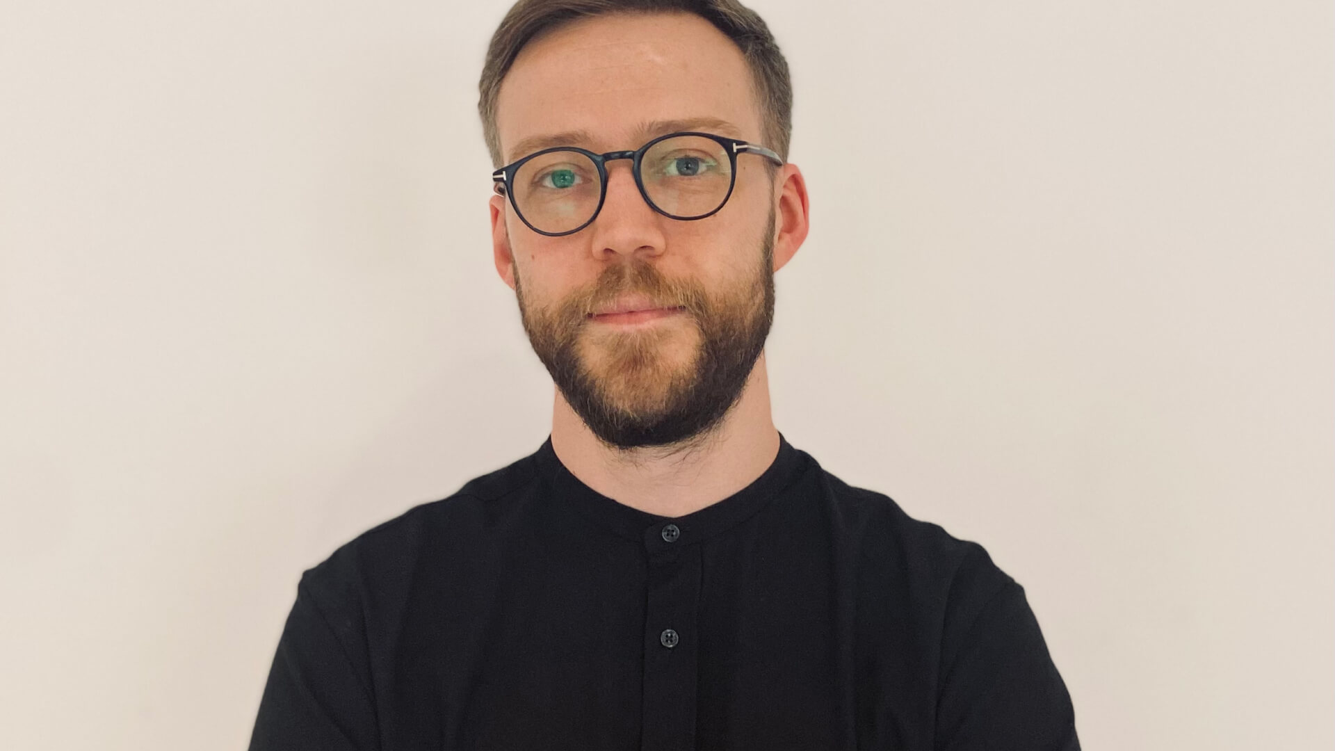 Man with a beard and glasses wearing a black buttoned shirt against a plain light background.