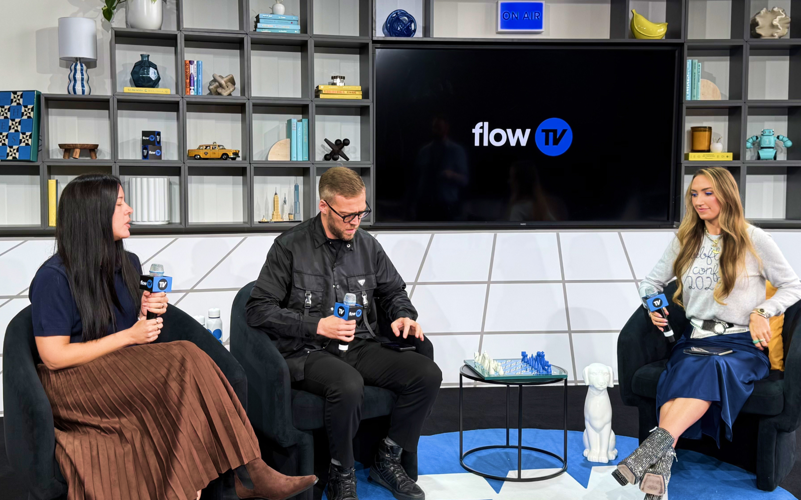 Three people seated on black chairs hold flow TV microphones on a set with chessboard and shelves behind.