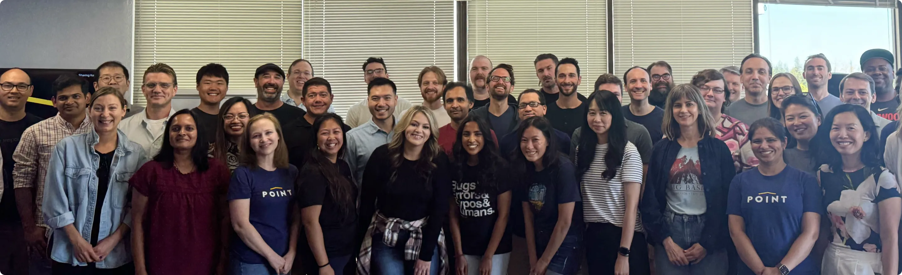 Large diverse group of office workers smiling and posing indoors in front of windows with blinds.