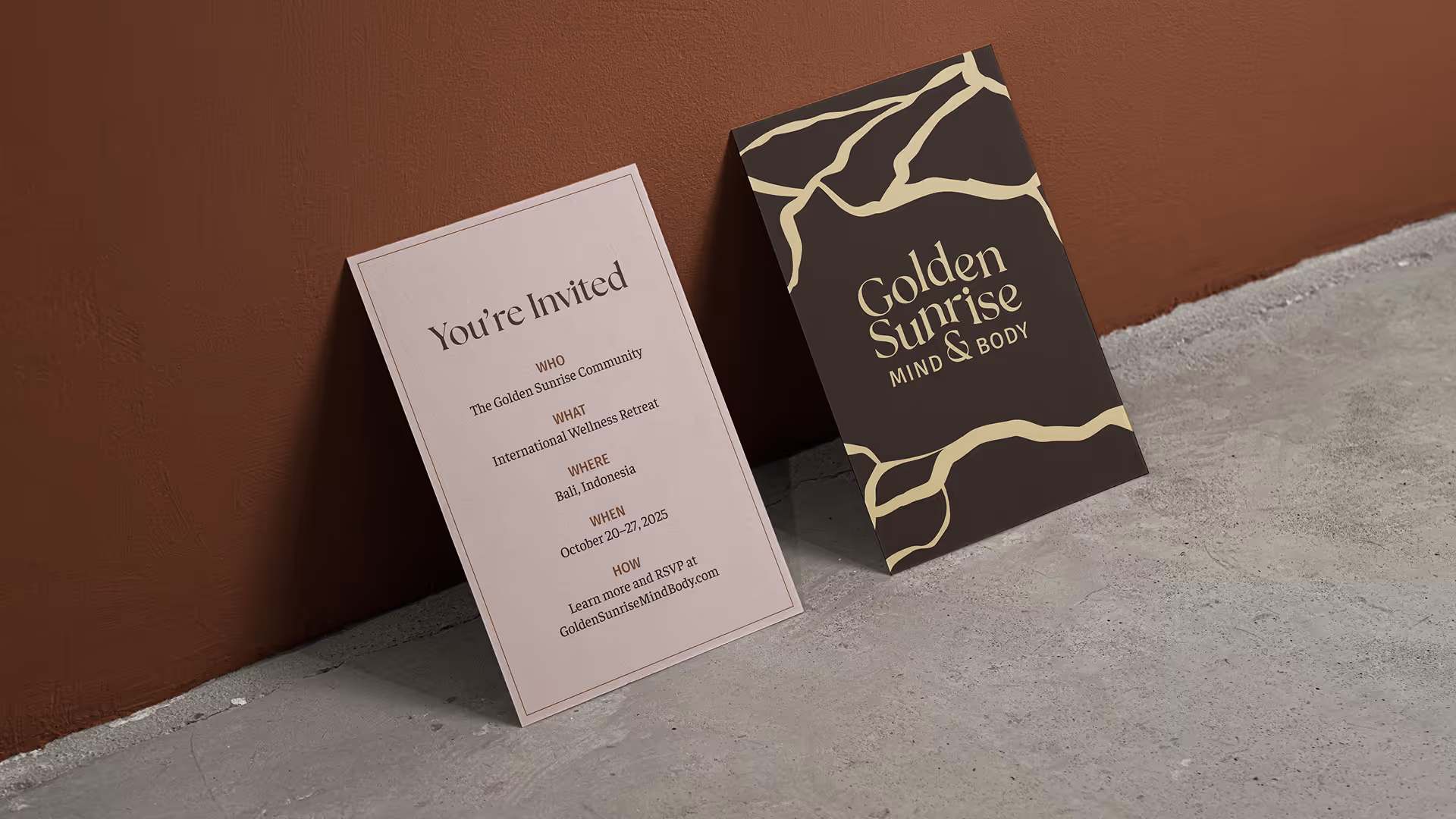 Mockup shows the front and back of an invitation printed on cardstock sitting on a taupe concrete floor and leaning against a terracotta-painted wall. The front heading reads "You're Invited" on a blush pink background. Body text reads "WHO The Golden Sunrise Community WHAT International Wellness Retreat WHERE Bali, Indonesia WHEN October 20–27, 2025 HOW Learn more and RSVP at GoldenSunriseMindBody.com". The back shows the Golden Sunrise logo framed by the brand pattern's golden kintsugi cracks against a raw umber background.