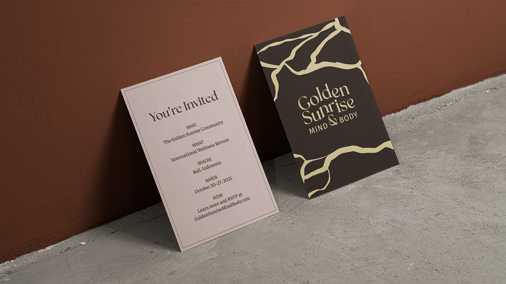 Mockup shows the front and back of an invitation printed on cardstock  sitting on a taupe concrete floor and leaning against a terracotta-painted wall. The front heading reads "You're Invited" on a blush pink background. Body text reads "WHO The Golden Sunrise Community WHAT International Wellness Retreat WHERE Bali, Indonesia WHEN October 20–27, 2025 HOW Learn more and RSVP at GoldenSunriseMindBody.com". The back shows the Golden Sunrise logo framed by the brand pattern's golden kintsugi cracks against a raw umber background. 