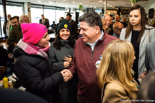 Anabel shaking hands with smiling Illinois governor J.B. Pritzker while wearing a hot pink winter hat embroidered with her campaign logo. Photo credit: Colin Boyle of Block Club Chicago.