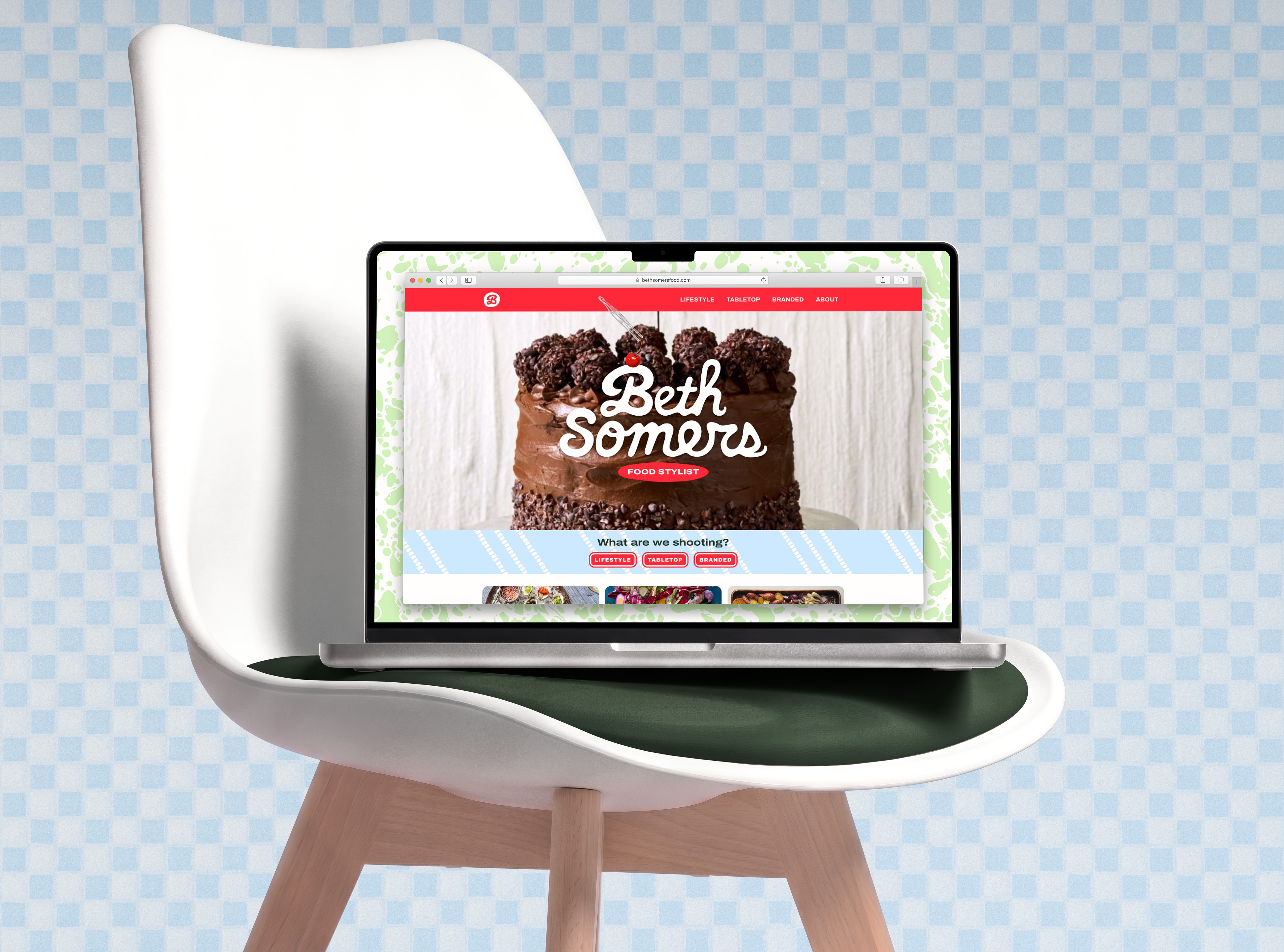 Mockup of the bethsomersfood.com landing page displayed in a Safari browser on a Macbook that rests on an Eames dining chair in front of a checkered tile wall. The desktop background is a mint-on-white enameled cookware pattern. The landing page hero section reads "Beth Somers: Food Stylist." Behind the text, we see one frame of a stop-motion video of a three-tiered chocolate cake being drizzled with chocolate sauce. Below this, a heading reads "What are we shooting?" followed by buttons labeled Lifestyle, Tabletop, and Branded.