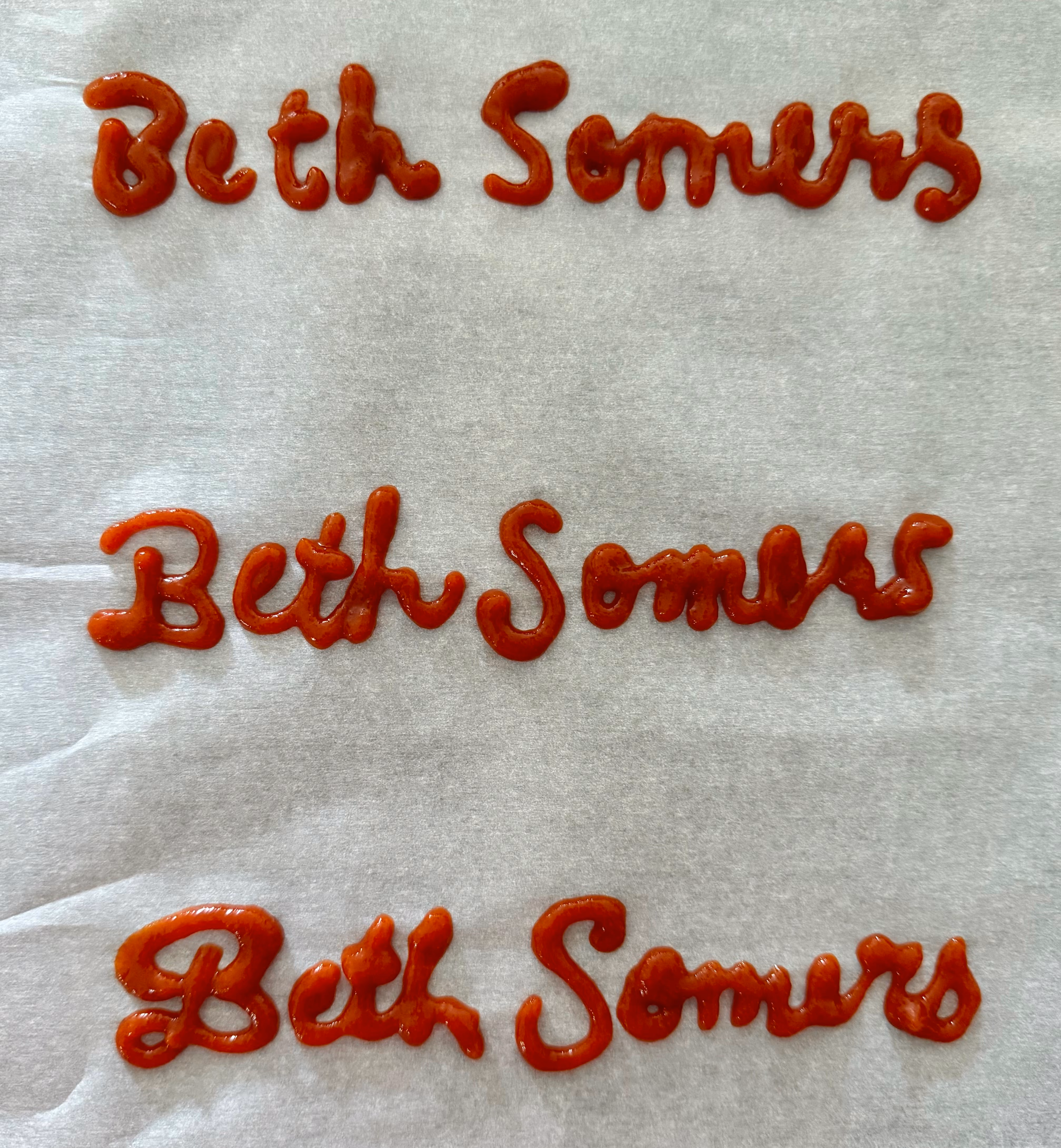 Three iterations of the Beth Somers logo handwritten with sriracha sauce on a sheet of parchment paper. The third iteration here served as the basis for the final saucy script logo.