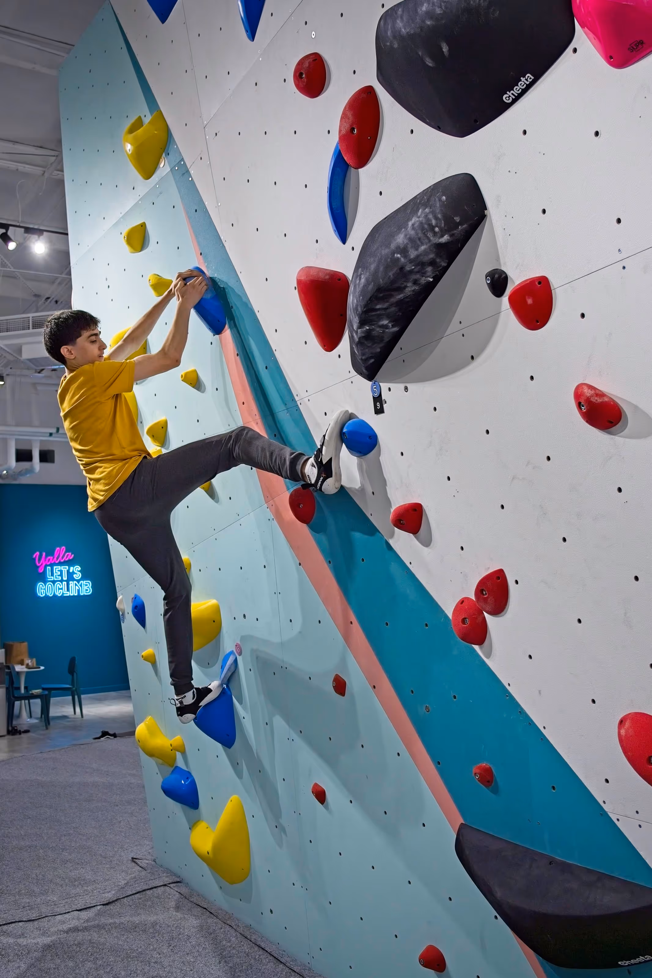 A young teenager bouldering. He is grabbing onto a big, blue climbing hand hold with his right foot reaching out to a big foothold on his right.