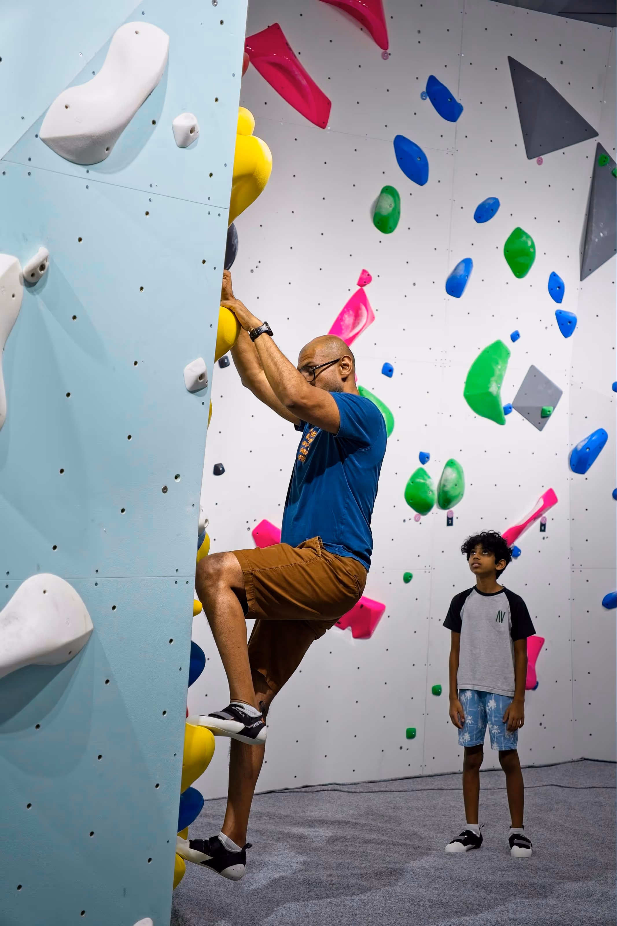 A young boy looks on as his father attempts bouldering.