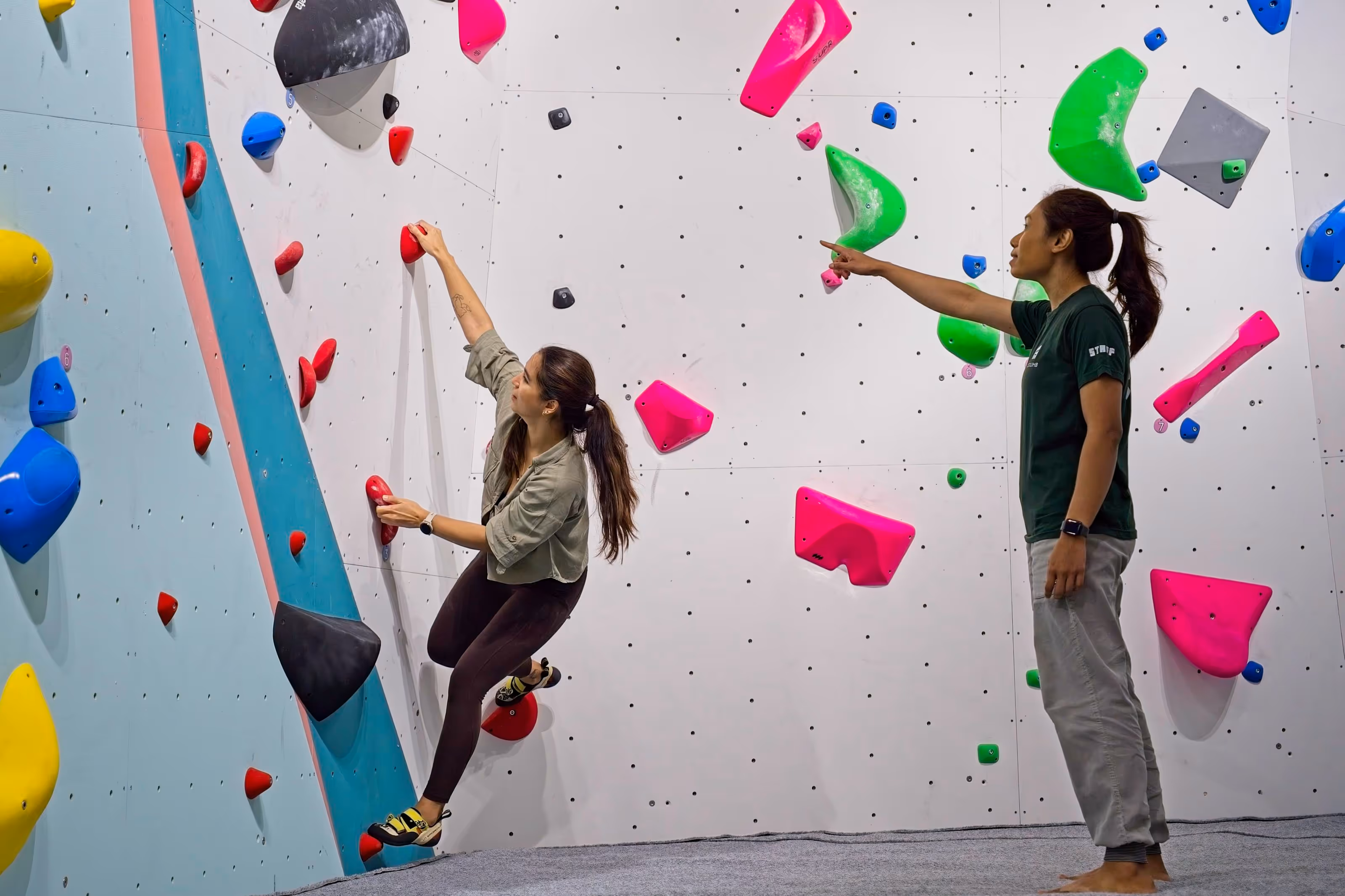 A lady bouldering while her female coach guides her along.