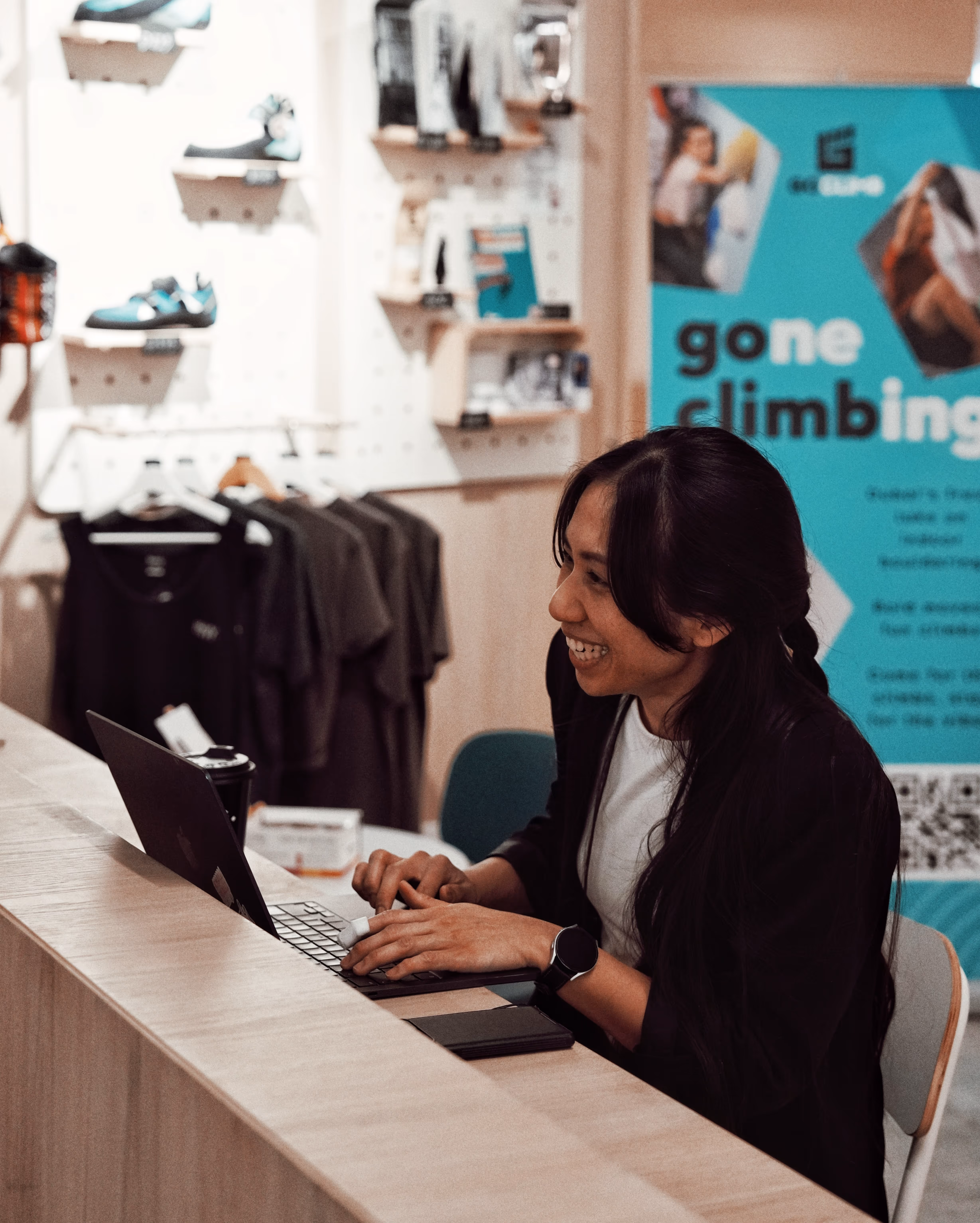Smiling woman typing on a laptop at a wooden counter in a store with climbing gear and clothing in the background.