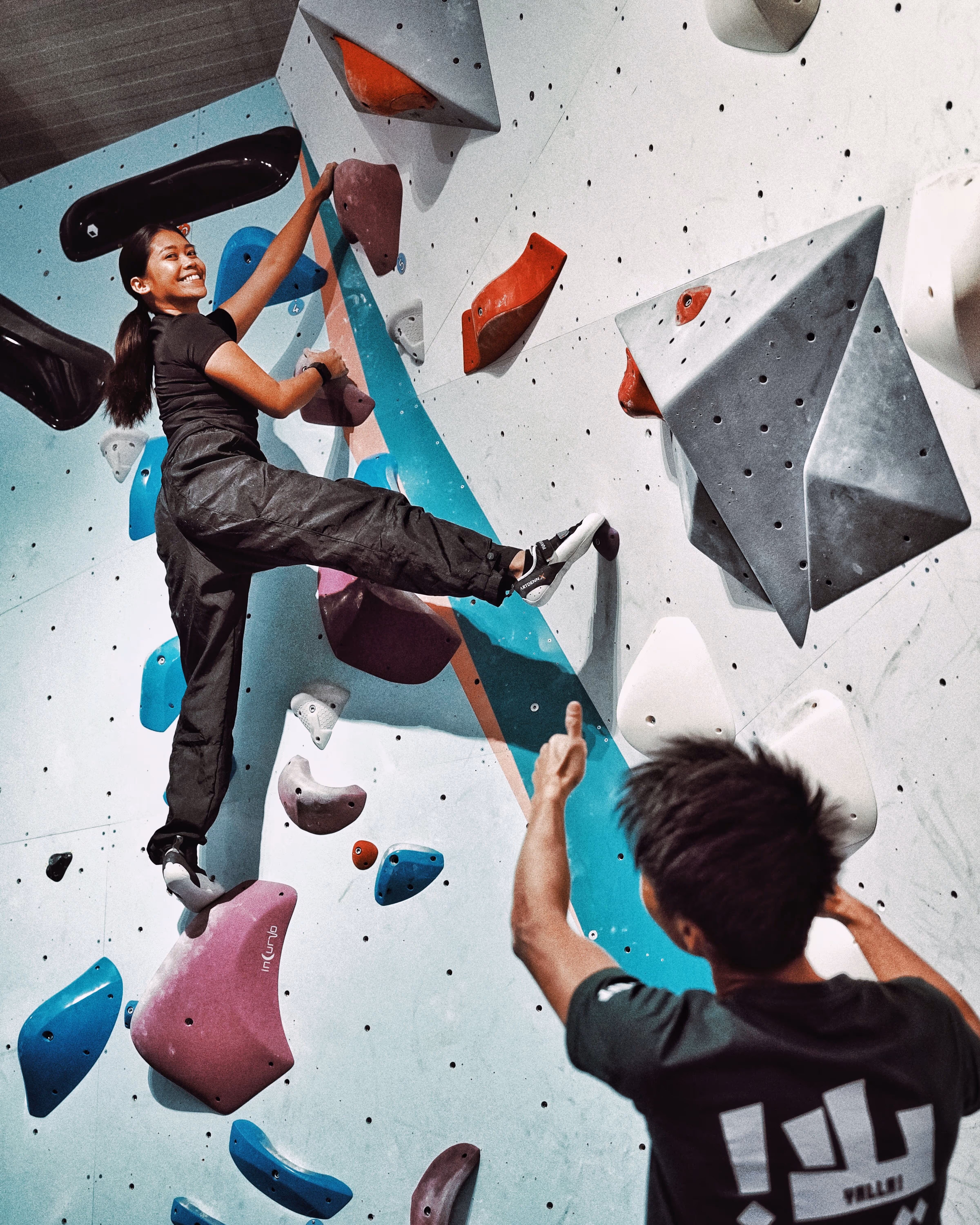 Smiling woman climbing an indoor bouldering wall with a coach giving her the thumbs up from below.