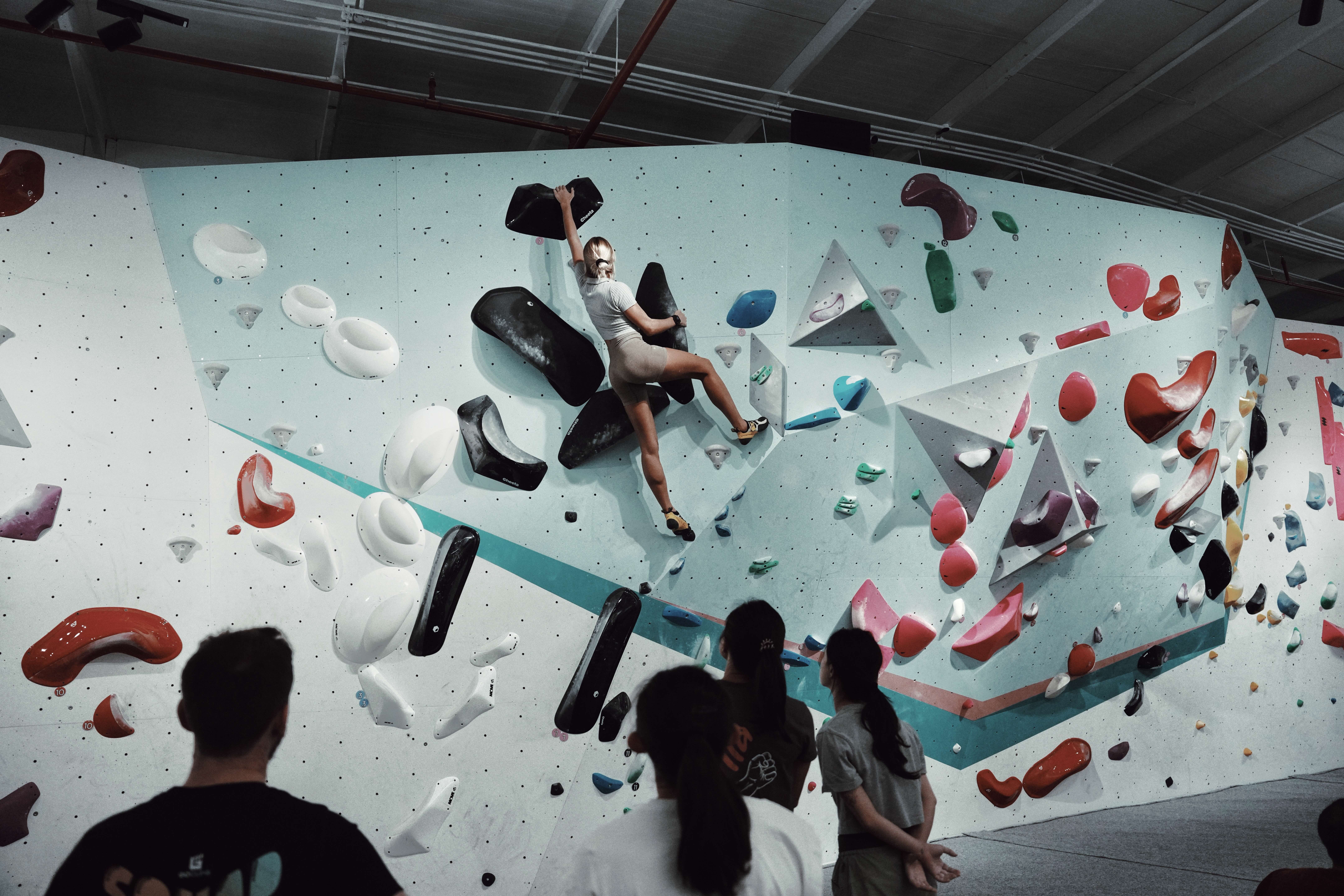 A woman climbing an indoor bouldering wall with a group watching her from below.
